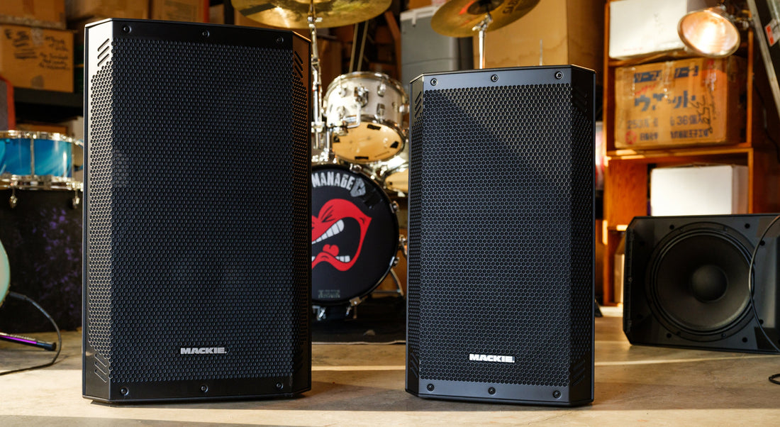 Mackie Thrash15v2 and Thrash12v2 powered PA speakers standing side by side in a rehearsal room with a drum kit behind them